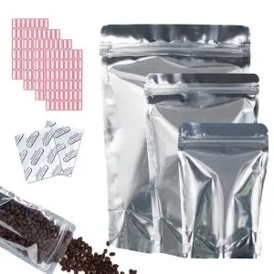 Três sacos Mylar prateados vazios da KanzoPack de diferentes tamanhos organizados ao lado de grãos de café, absorvedores de oxigênio e etiquetas.
