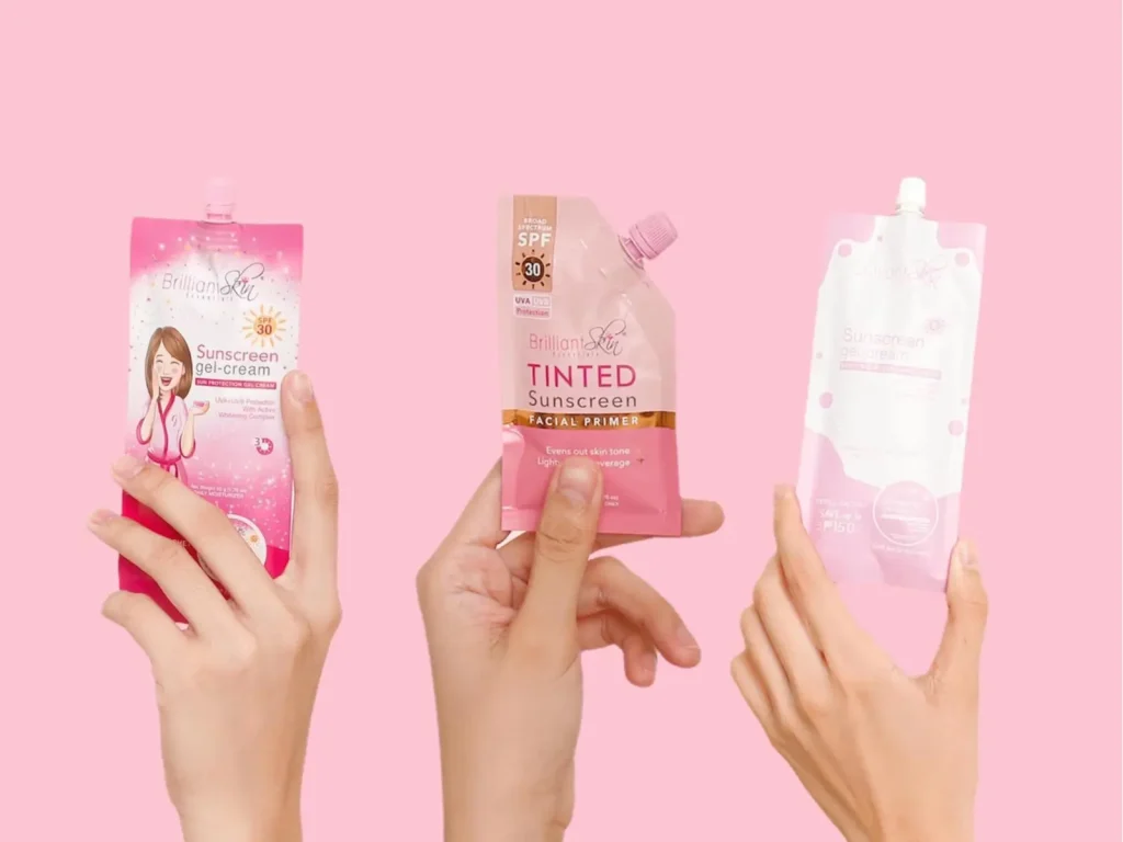 Três mãos segurando diferentes sachês de protetor solar e primer da Brilliant Skin Essentials contra um fundo rosa.