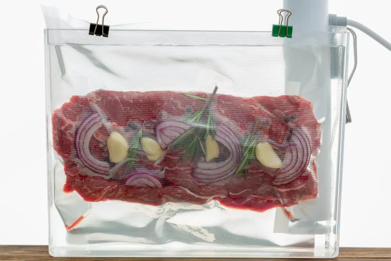 Un filete crudo sazonado con cebolla roja, ajo y romero dentro de una bolsa sellada al vacío sumergida en un baño de agua para cocción al vacío.